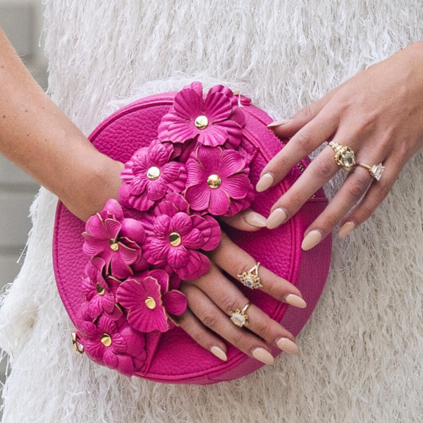 A pink handbag with a 3D floral applique design, gold-tone studs, and a pebbled vegan leather texture. It is shown being held by a person with a white background.