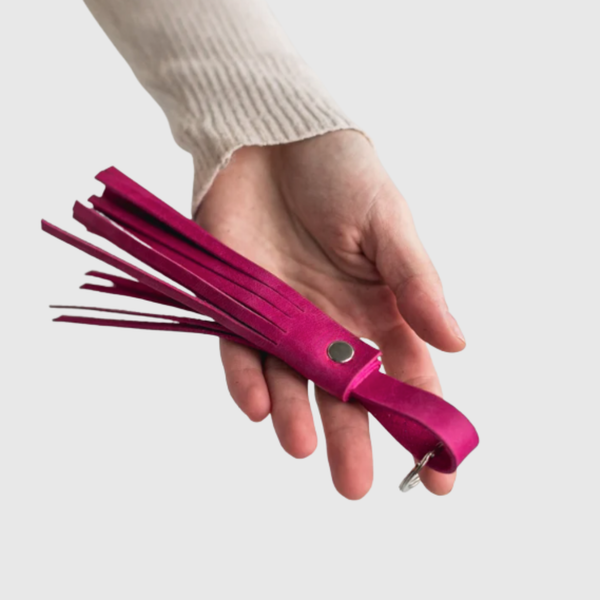 Hand holding a pink leather flogger against a light background